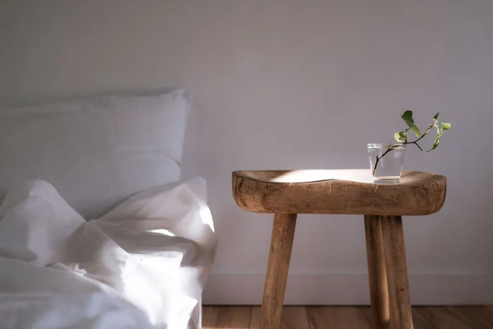 A wooden stool with a single stem in a vase beside an unmade linen bed